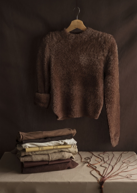 A brown textured sweater hangs above a neatly folded stack of neutral‑toned clothing, set against a dark backdrop with arranged twigs, creating a warm, monochromatic scene that highlights fabric texture and materiality.