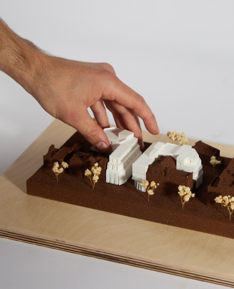 A small site model on a brown base with white structures, touched by a hand.
