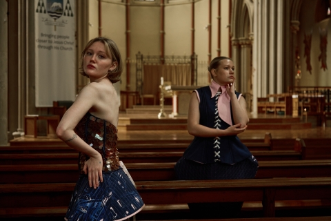 two models posing in a church setting standing between the pews. they are both wearing blue garments
