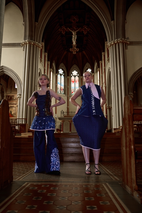 two models in blue garments standing in aisle of what looks like a church facing forwards