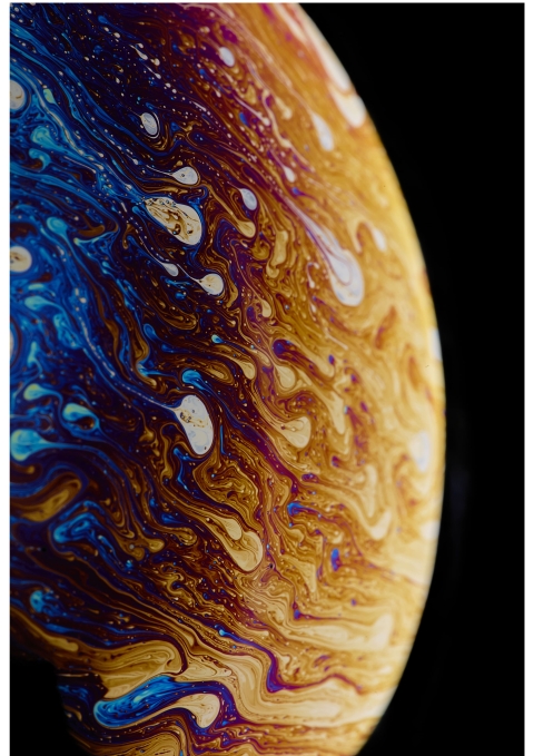 A close‑up of swirling blue, gold, purple, and white patterns on a soap‑bubble surface, creating an abstract, marbled effect.
