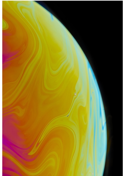 A close‑up of swirling yellow, orange, pink, and blue patterns on a soap‑bubble surface against a black background.