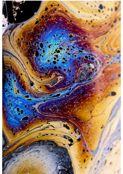 A close‑up of swirling blue, purple, orange, yellow, and black fluid patterns with small dark specks, creating a marbled, liquid‑like abstract surface.